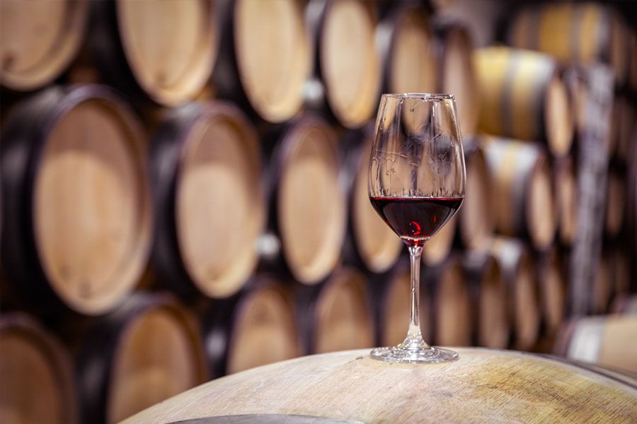 Closeup red wine glass on background of wooden oak barrels stacked in straight rows in order in cellar of winery, vault. Concept professional degustation, winelover, sommelier travel