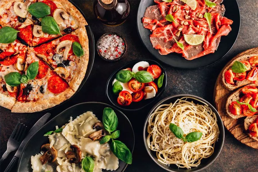 Full table of italian meals on plates Pizza, pasta, ravioli, carpaccio. caprese salad and tomato bruschetta on black background. Top view